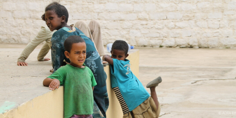 Zwei Kinder stehen vor einer Mauer; eines hebt das Bein, das andere klettert hoch, während oben schon ein Kind sitzt