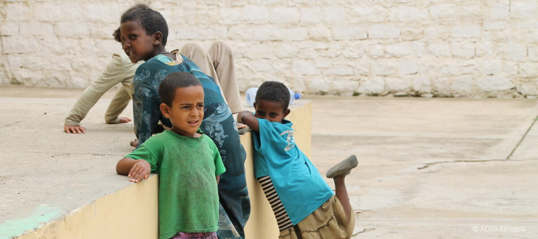 Zwei Kinder stehen vor einer Mauer; eines hebt das Bein, das andere klettert hoch, während oben schon ein Kind sitzt