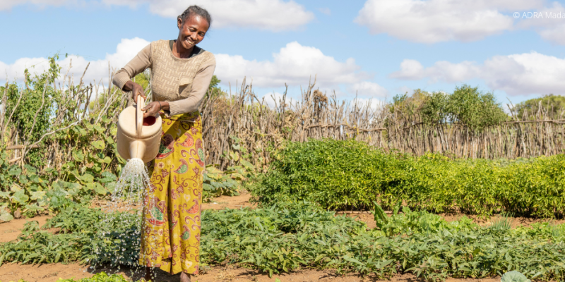 Eine lachende Frau hält mit beiden Händen eine Gießkanne und bewässert ihre Bio Ernte.