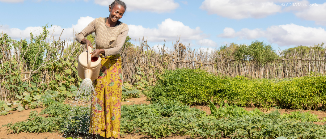 Eine lachende Frau hält mit beiden Händen eine Gießkanne und bewässert ihre Bio Ernte.