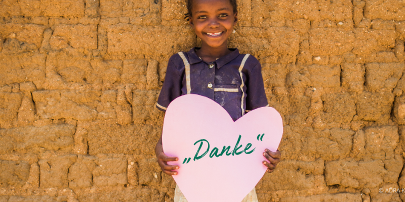 Ein Mädchen aus Kenia hält ein rosa Herz aus Papier mit beiden Händen, in welchem mit einem schrägen Schriftzug "Danke" zu lesen ist..