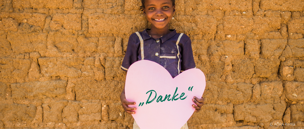 Ein Mädchen aus Kenia hält ein rosa Herz aus Papier mit beiden Händen, in welchem mit einem schrägen Schriftzug "Danke" zu lesen ist..