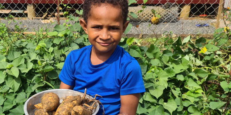 Ein junges Kind hält eine Schüssel mit Pflanzenwurzeln in beiden Händen und lächelt in die Kamera.