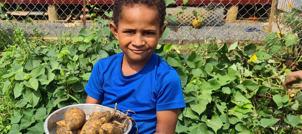 Ein junges Kind hält eine Schüssel mit Pflanzenwurzeln in beiden Händen und lächelt in die Kamera.