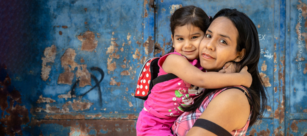 Guter Start ins Leben: Lächelnde Mutter hält fröhlich lächelnde Tochter im Arm, beide blicken in die Kamera.