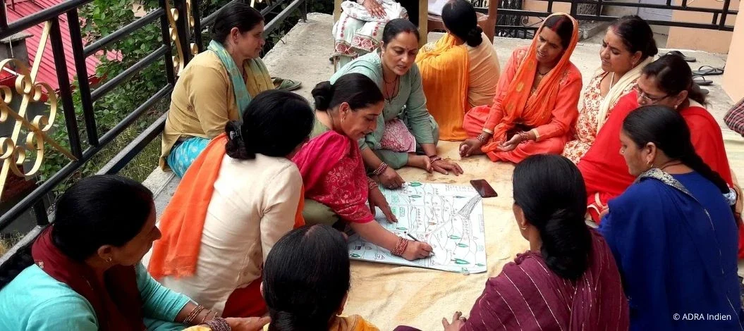 Eine Gruppe von Frauen sitzt im Kreis, diskutiert und arbeitet gemeinsam an einem großen Papierbogen mit Zeichnungen und Plänen auf einer Terrasse in Indien