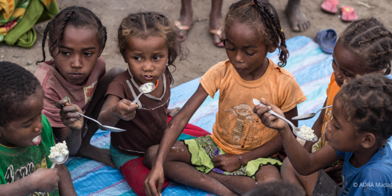 Eine Gruppe Kinder und Kleinkinder sitzt um eine Schüssel mit Brei gefüllt und teilt sich das Essen