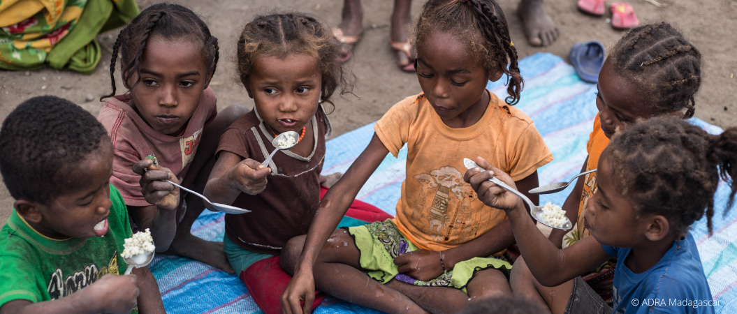Eine Gruppe hungernde Kinder und Kleinkinder sitzt um eine Schüssel mit Brei gefüllt und teilt sich das Essen