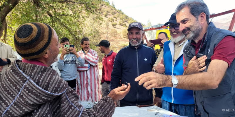 ADRA-Mitarbeiter sprechen herzlich mit einem älteren Dorfbewohner in Marokko, umgeben von lachenden Menschen.
