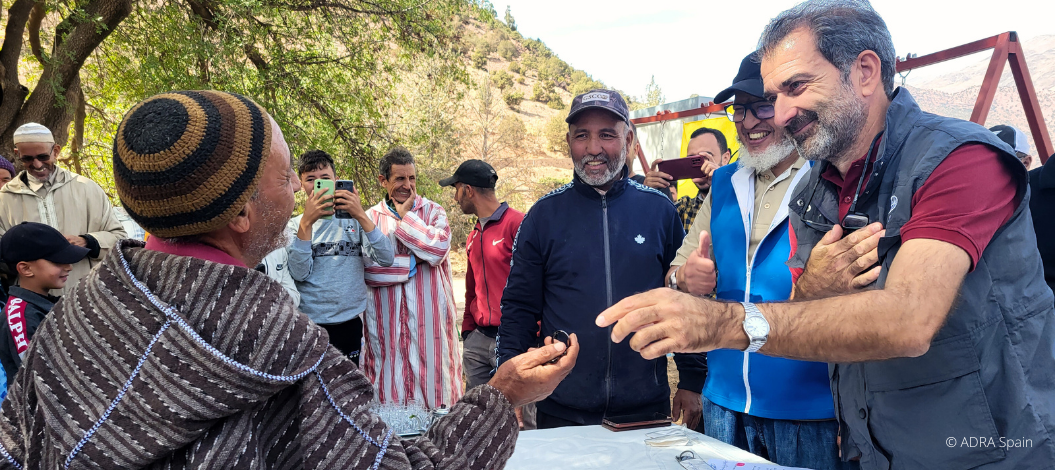 ADRA-Mitarbeiter sprechen herzlich mit einem älteren Dorfbewohner in Marokko, umgeben von lachenden Menschen.
