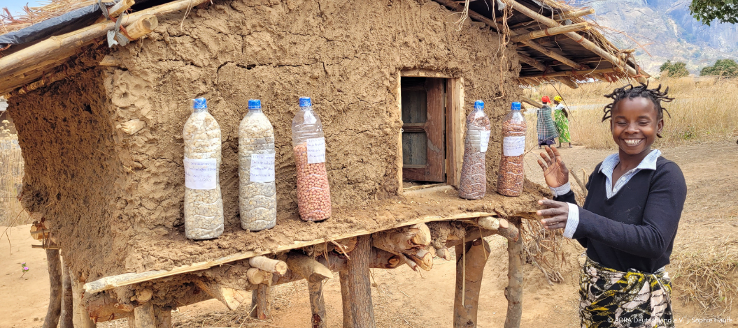 Ein Mädchen steht vor einer Saatgutbank in Mosambik, die mit Plastikflaschen voller lokalem Saatgut gefüllt ist. Sie lächelt stolz und präsentiert die Saatgutsorten, die für die nachhaltige Landwirtschaft und Ernährungssicherheit in der Region von entscheidender Bedeutung sind. Diese Saatbank ist Teil eines Projekts von ADRA, das Frauen und Landwirt:innen mit technischem Wissen und Ressourcen unterstützt, um die landwirtschaftliche Produktivität und die Widerstandsfähigkeit gegenüber dem Klimawandel zu steigern.