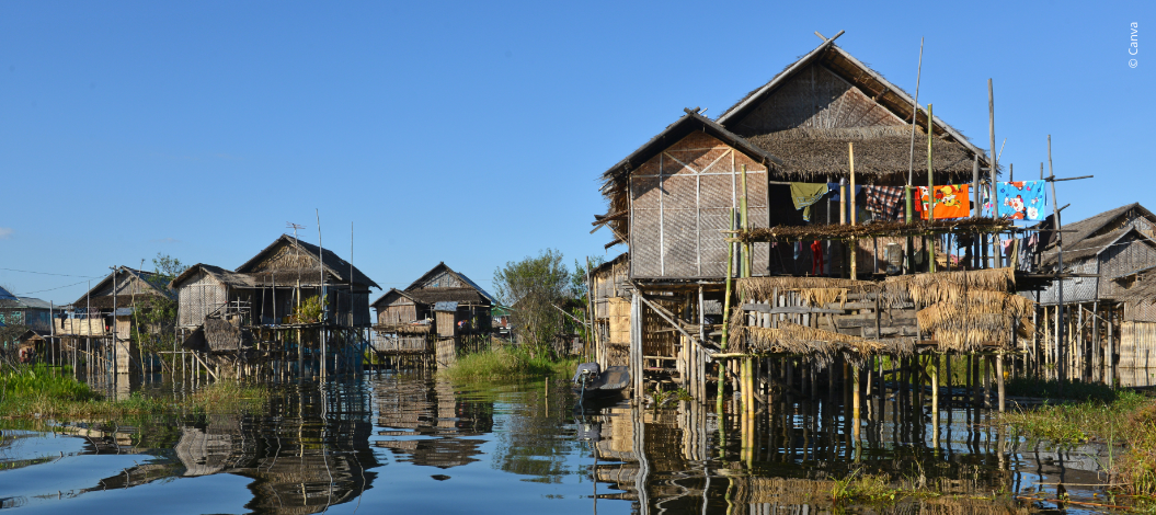 Traditionelle Pfahlbauten in Myanmar aus Bambus und Holz spiegeln sich im Fluss – mit Wäscheleinen an den Balkonen