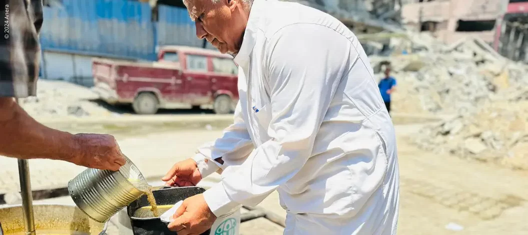 Gaza Nothilfe - Älterer Mann erhält Suppe aus großem Topf mit ADRA-Logo – im Hintergrund Trümmer und zerstörte Gebäude