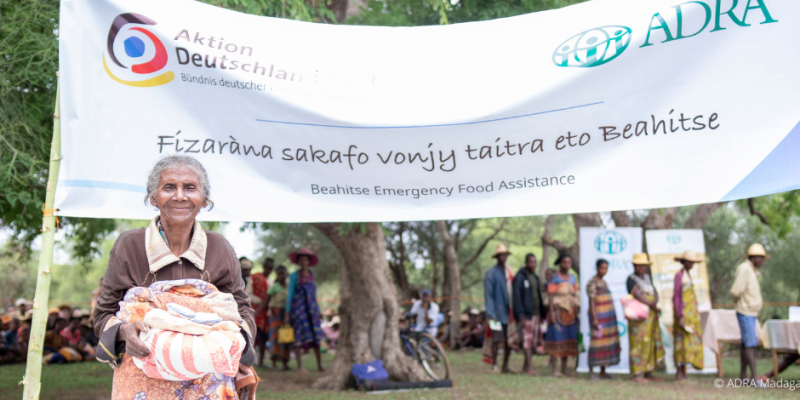 Ältere Frau in Madagaskar hält Hilfsgüter vor ADRA- und ADH-Banner, hinten warten Menschen in der Schlange