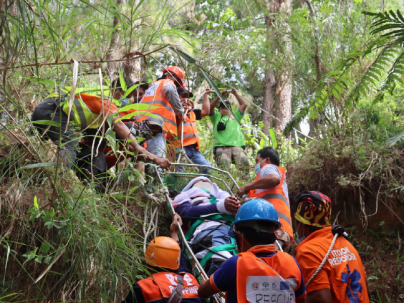 Ein Hilfstrupp im Wald transportiert einen Verletzen Menschen einen Hang zwischen den Bäumen hoch