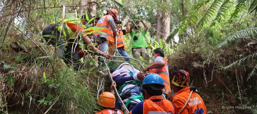 Ein Hilfstrupp im Wald transportiert einen Verletzen Menschen einen Hang zwischen den Bäumen hoch
