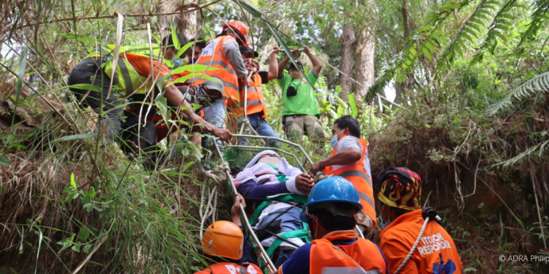 Ein Hilfstrupp im Wald transportiert einen Verletzen Menschen einen Hang zwischen den Bäumen hoch