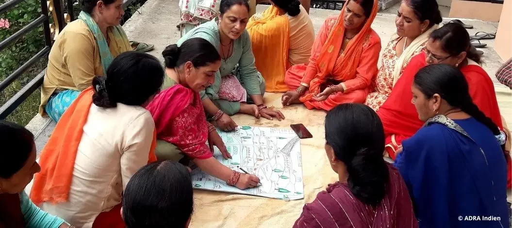 Eine Gruppe von Frauen aus Indien sitzt im Kreis auf dem Boden und diskutiert über ein großes Blatt Papier mit Diagrammen. Sie scheinen gemeinsam an einer Planung oder Schulung teilzunehmen.