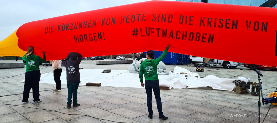 ADRA-Team hält rotes Luftobjekt mit dem Slogan „Die Kürzungen von heute sind die Krisen von morgen! #luftnachoben“