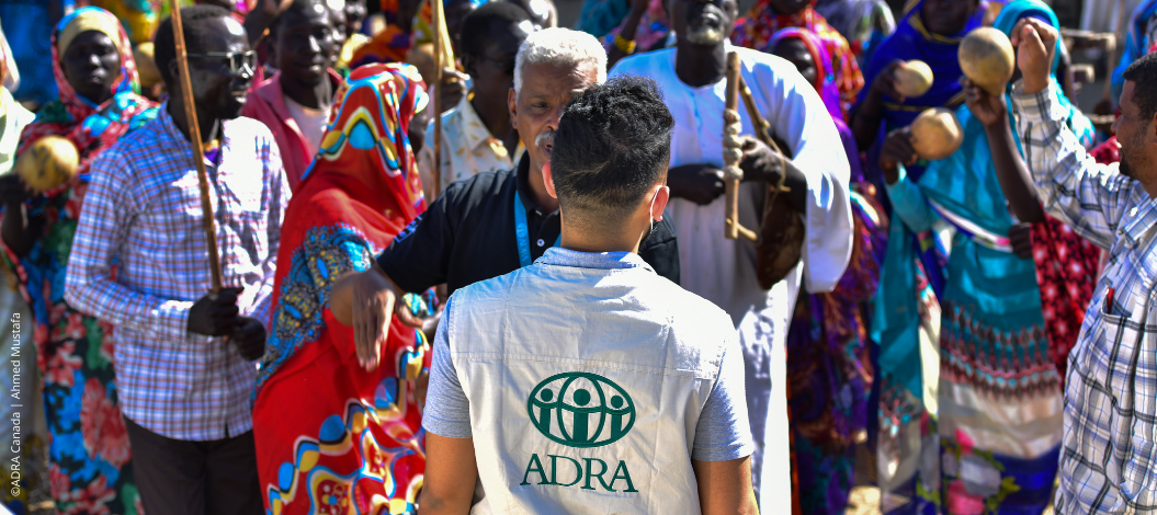 ADRA-Mitarbeiter steht vor bunt gekleideter Menschenmenge in Afrika – Versammlung mit Musik und Gesprächen im Freien