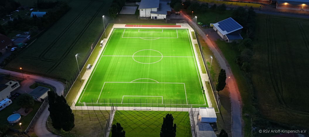 Neuer Kunstrasenplatz abends beleuchtet durch Flutlichtanlage, mit Blick auf das neue Kleinspielfeld. Drohnenaufnahme vom RSV Arloff-Kirspenich e.V.