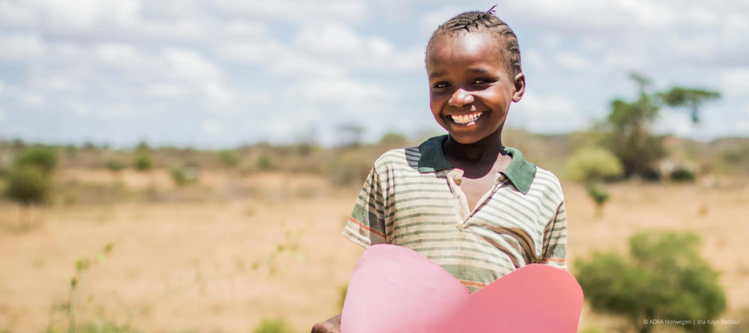 Lächelndes Kind in Kenia hält ein großes rotes Papierherz mit beiden Händen vor sich.