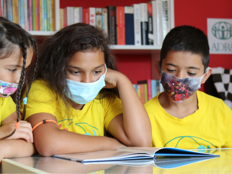 Drei Grundschulkinder in gelben ADRA-TShirts sitzen mit Mundschutz an einem Tisch und lernen