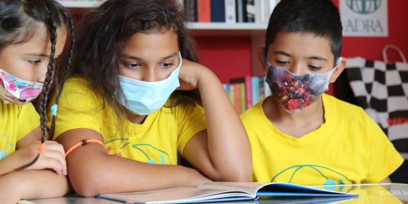 Drei Grundschulkinder in gelben ADRA-TShirts sitzen mit Mundschutz an einem Tisch und lernen
