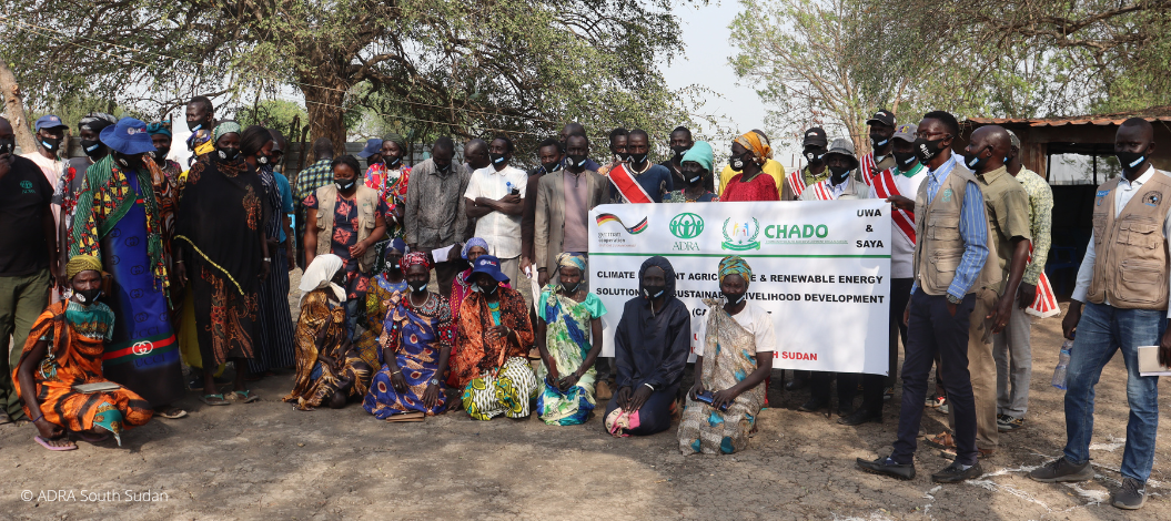 Eine Gruppe aus mehr als 20 Menschen, die im Südsudan Hilfsgelder von ADRA empfangen hat, sitzt vor Bäumen und stellt mit einem Schild das Projekt von ADRA vor