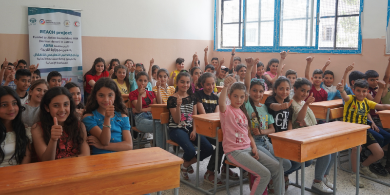 Eine gemischt Schulklasse sitzt in einem Klassenzimmer. Alle Kinder strecken die Hände nach oben und strahlen Freude aus.