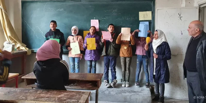 Sieben Schüler:innen stehen vor der Tafel und halten jeweils ein farbiges Papier hoch, auf den ein Buchstabe abgebildet ist