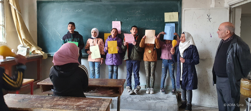 Sieben Schüler:innen stehen vor der Tafel und halten jeweils ein farbiges Papier hoch, auf den ein Buchstabe abgebildet ist