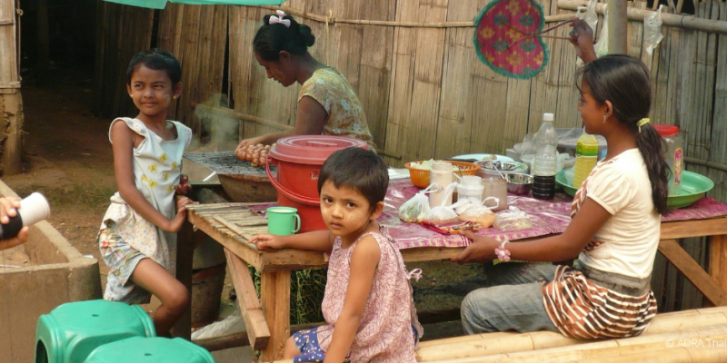 Kinder sitzen am Tisch und essen, eine Frau kocht