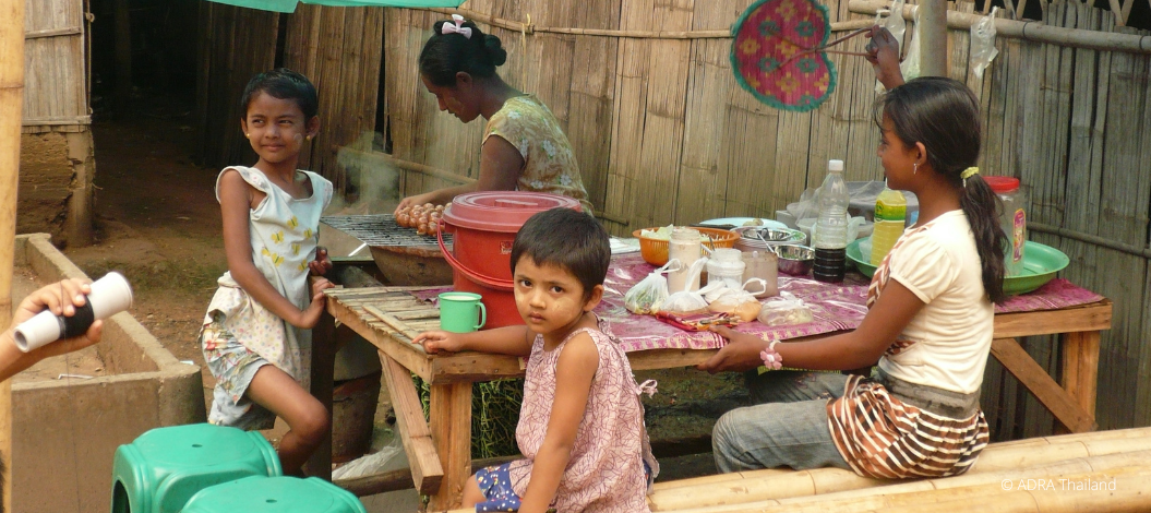 Kinder sitzen am Tisch und essen, eine Frau kocht