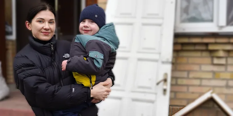 Eine junge Mutter aus der Ukraine hält liebevoll ihren Sohn im Arm, während sie vor einem zerstörten Haus steht, an dessen Wand die Tür gegen die Wand angelehnt ist. Sie benötigen Winterhilfe