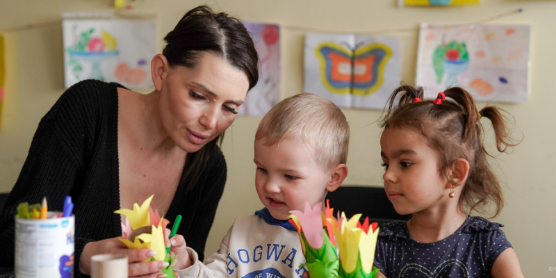 Eine Frau zur Linken sitzt mit zwei Kindern am Tisch und malt mit ihnen.