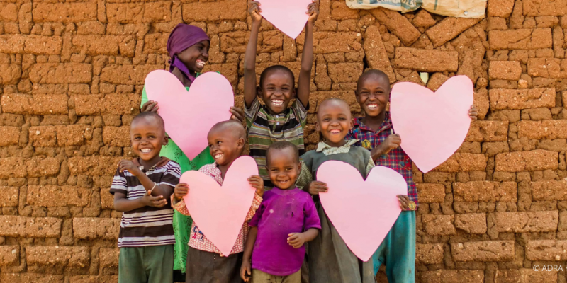 Lachende Kinder in Kenia halten große rosa Herzen vor einer Mauer aus Lehmziegeln
