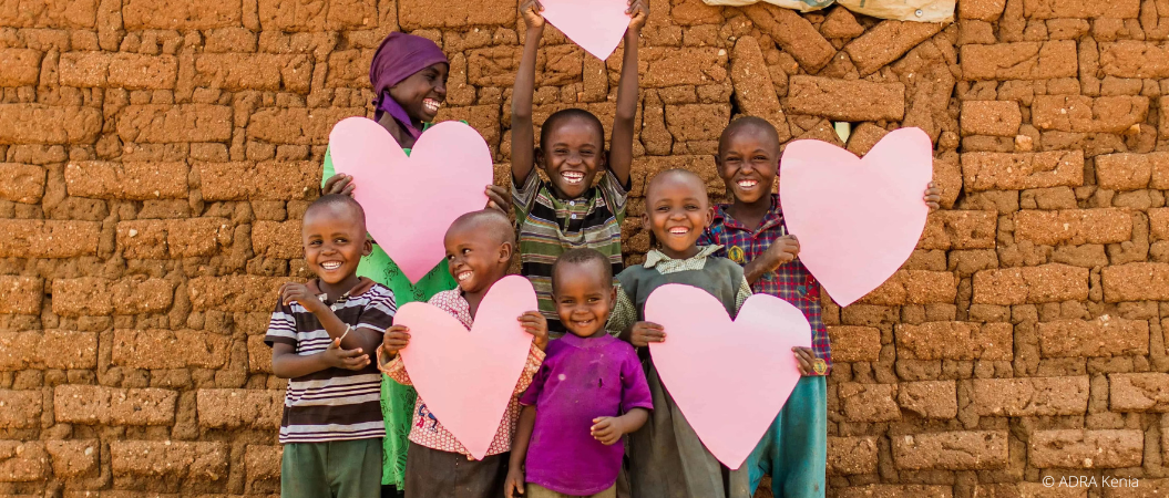 Lachende Kinder in Kenia halten große rosa Herzen vor einer Mauer aus Lehmziegeln