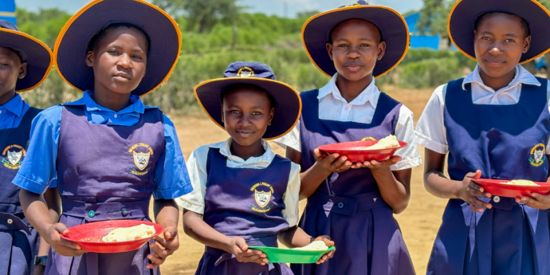 Eine junge Schulklasse aus Zimbabwe steht im Freien und hält Teller mit Essen in den Händen, während sie in die Kamera lächeln.