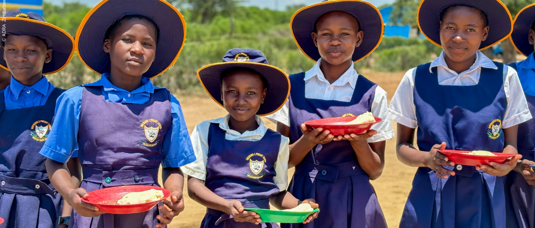 Eine junge Schulklasse aus Zimbabwe steht im Freien und hält Teller mit Essen in den Händen, während sie in die Kamera lächeln.