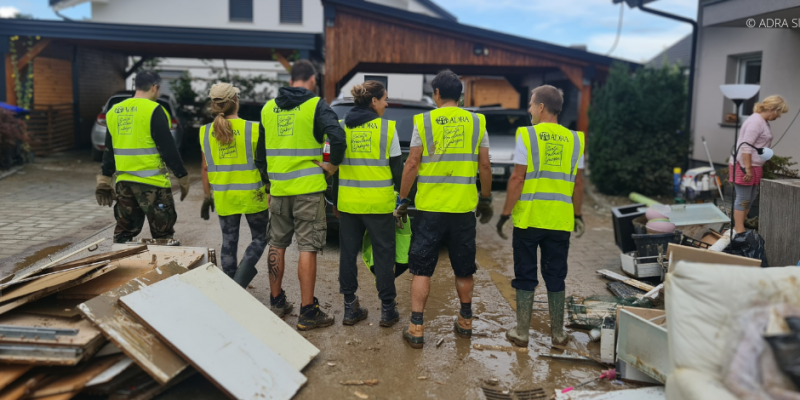 Sechs ADRA-Helfer in gelben Westen unterstützen nach der Flut in Slowenien, umgeben von zerstörten Möbeln und Gegenständen