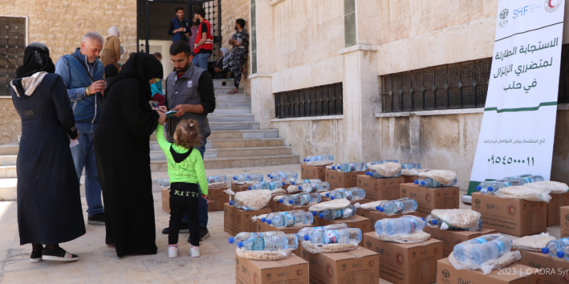 Bereitgestellte Lebensmittelpakete vor einem ADRA-Roll-Up-Plakat für die Erdbebenhilfe in Syrien, während Menschen auf humanitäre Unterstützung warten.