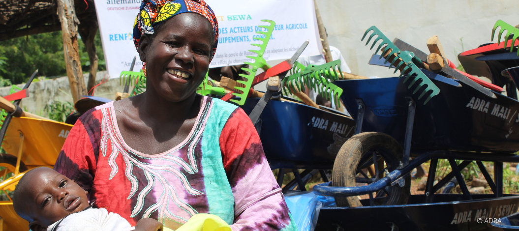 Frau mit Kind im Arm lächelt in die Kamera, hinter ihr Gartenwerkzeug und ein ADRA-Schild