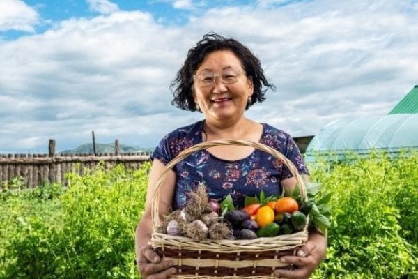 Eine Frau steht mit einem gut gefüllten Gemüsekorb lächelnd vor einem Gewächshaus auf einem Feld