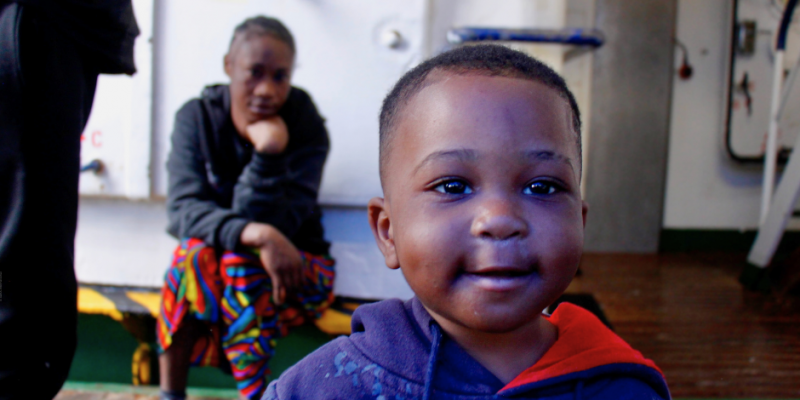 Kleiner Junge blickt zufrieden in die Kamera, Mutter im Hintergrund an Bord des Rettungsschiffs ‚Humanity 1‘ von SOS Humanity