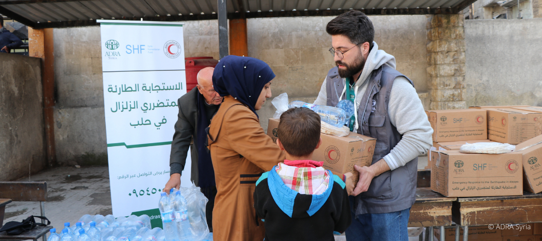 Notfallhilfe nach schwerem Erdbeben in Nord-Syrien: ADRA-Mitarbeiter übergibt Nothilfe-Pakete an eine Frau und ihr Kind. Im Hintergrund hält ein Mann Wasserflaschen bereit.