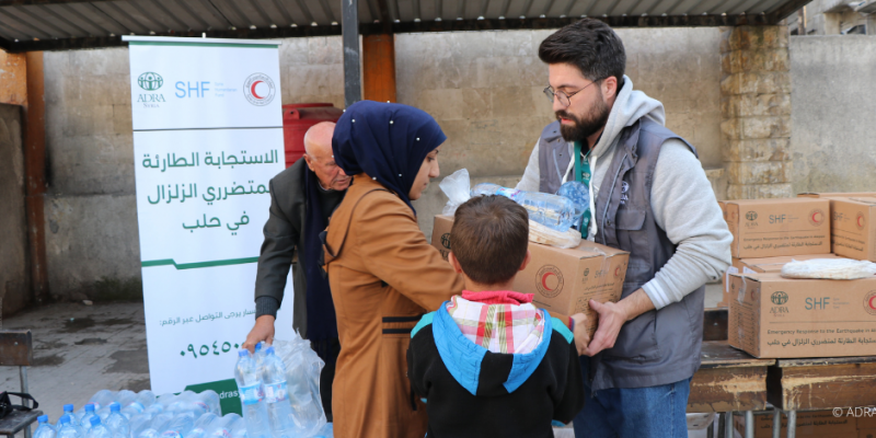 Nach Erdbeben in Nordsyrien: ADRA-Mitarbeiter übergibt Nothilfe-Pakete an Frau mit Kind, Mann hält Wasserflaschen