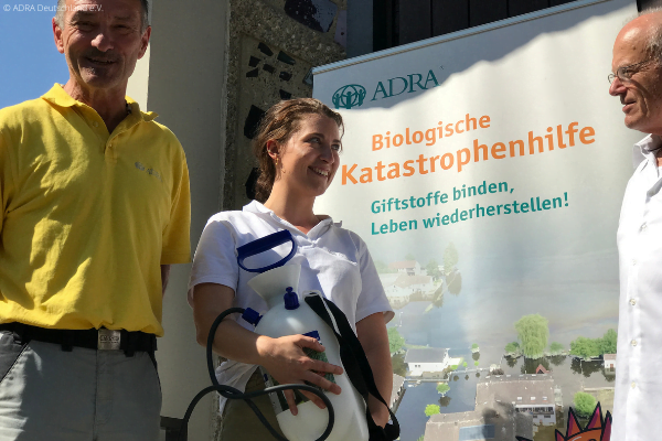 Eine lächelnde Frau steht zwischen zwei Männern, vor einem ADRA-Plakat und hält eine Pumpe für effektive Mikroorganismen in den Händen