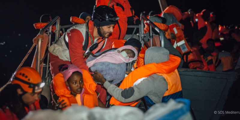 Helfer übergibt gerettetes Kleinkind an Eltern auf Booten im Mittelmeer – Teil der Mission von SOS Mediterranee