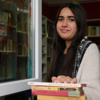 Eine Schülerin steht in einem Raum mit Bücherregalen lächelnd vor einem offen Fenster, auf dessen Fensterbank vier Bücher liegen, auf welche die Hand des Mädchens draufliegt